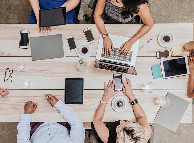 Group of people sitting around a desk discussing projects while using computers and other devices